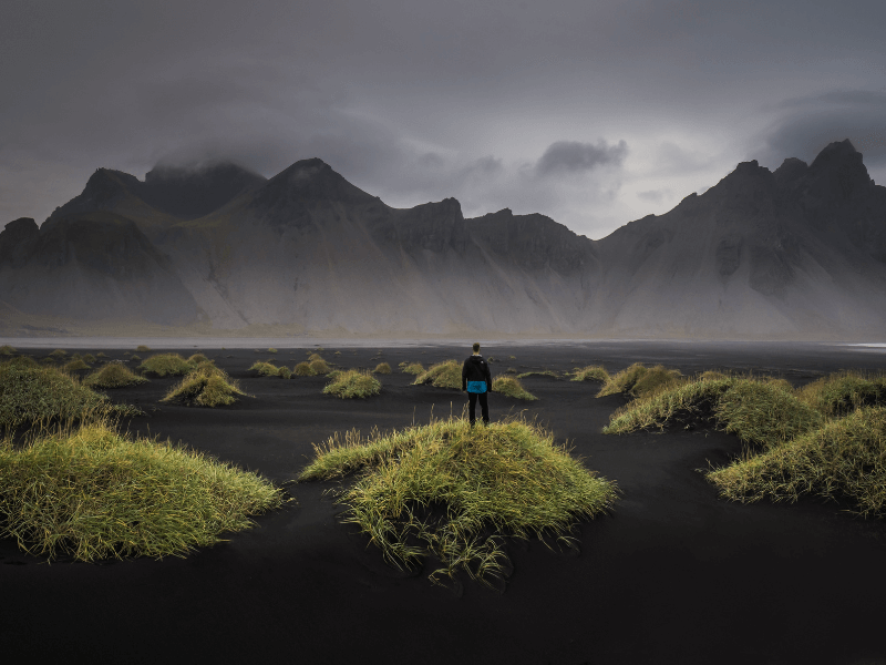 Persoon staat op een grasheuvel met uitzicht op een zwart zandstrand en imposante bergen gehuld in mist, IJsland.