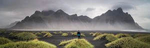 Persoon staat op een grasheuvel met uitzicht op een zwart zandstrand en imposante bergen gehuld in mist, IJsland.