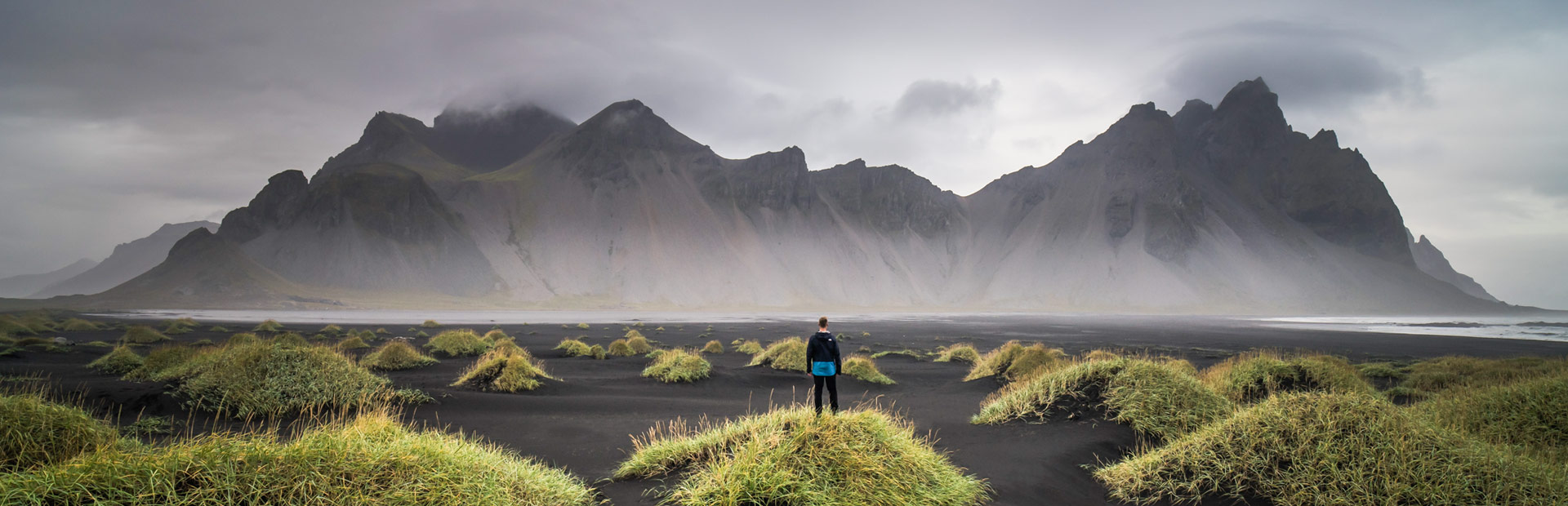 Persoon staat op een grasheuvel met uitzicht op een zwart zandstrand en imposante bergen gehuld in mist, IJsland.