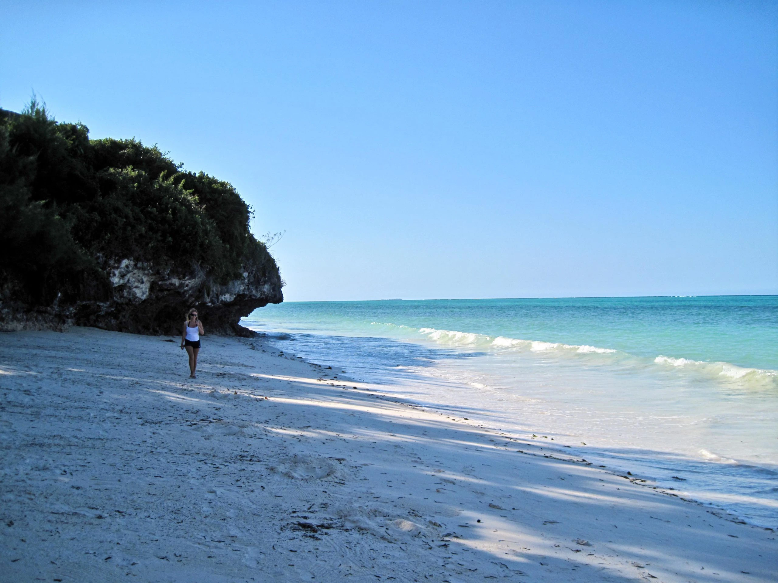 Rustig strand met wit zand en turquoise zee op Zanzibar, Tanzania, met een vrouw die langs de vloedlijn wandelt.