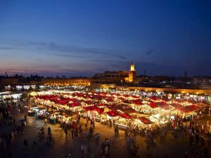 Avondbeeld van het drukke Djemaa el Fna-plein in Marrakesh, met verlichte marktkramen onder rode daken en mensen die rondlopen.