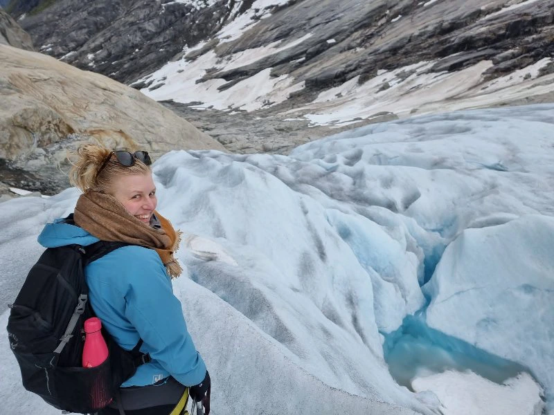 Vrouw met rugzak en warme kleding glimlacht terwijl ze op een gletsjer staat, omringd door ijs en besneeuwde rotsen.