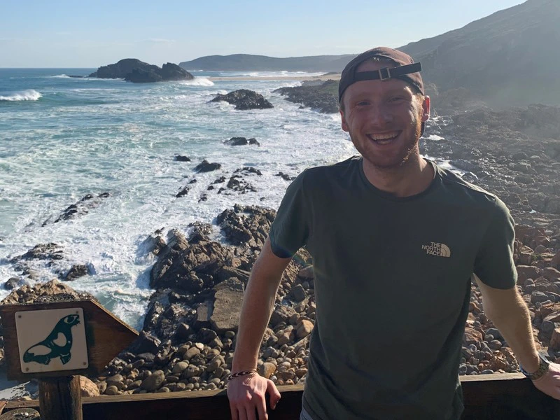 Lachende man staat bij een houten railing met uitzicht op een rotsachtige kustlijn en de oceaan, naast een bordje met een zeehond-symbool.