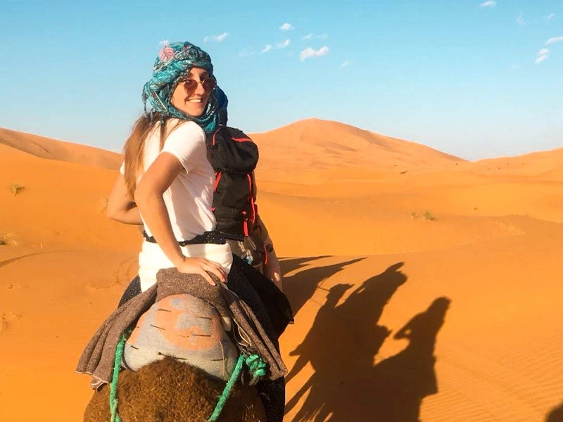 Reiziger rijdt op een kameel door de oranje zandduinen van de woestijn onder een blauwe lucht, met lange schaduwen op het zand
