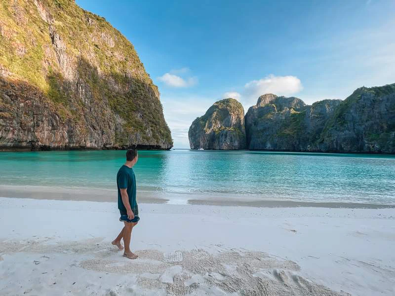 Man loopt op een wit zandstrand aan een turquoise baai, omringd door hoge groene rotskliffen onder een blauwe lucht met enkele wolken.