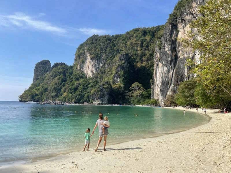 Vrouw met twee kinderen loopt langs een wit zandstrand met turquoise water, omringd door hoge groene kalksteenkliffen.