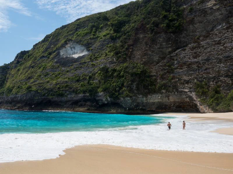 Twee mensen lopen langs een verlaten zandstrand met helderblauw water en een steile, groene klif op de achtergrond.
