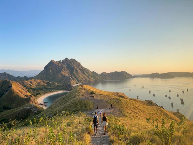 Wandelaars volgen een smal pad over een heuvelrug met uitzicht op een baai en grillige bergen tijdens zonsondergang bij