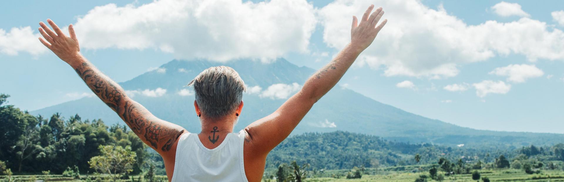Man met getatoeëerde armen en witte tanktop staat met opgeheven armen en kijkt uit over een groen landschap met een berg op de achtergrond onder een blauwe lucht met wolken.