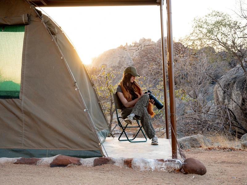 Tent met luifel in een droge, rotsachtige omgeving, waar een vrouw op een klapstoel zit met een camera in haar handen.