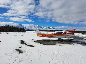 Klein propellervliegtuig met oranje en witte kleuren staat op een besneeuwde landingsbaan, met besneeuwde bergen en een blauwe lucht met wolken op de achtergrond.