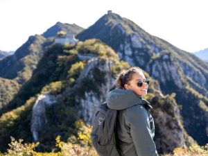 Vrouw met zonnebril en rugzak glimlacht terwijl ze wandelt in de bergen, met op de achtergrond groene bergtoppen en delen van de Chinese Muur.