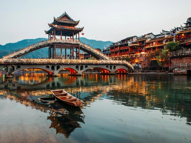 Traditionele boogbrug met paviljoen over een rivier in een Chinees stadje, met houten bootjes op het water en verlichte gebouwen langs de oever.