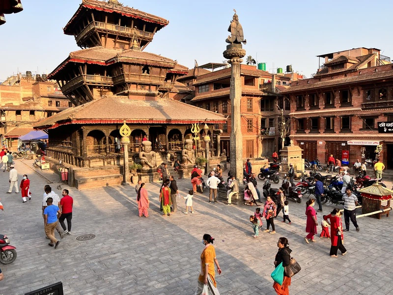 Drukke pleinscene in Bhaktapur, Nepal, met mensen die wandelen tussen historische tempels en traditionele roodbruine gebouwen onder een heldere hemel.