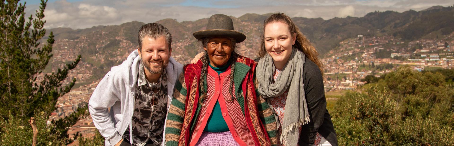 Twee reizigers poseren lachend naast een lokale vrouw in traditionele kleding, met bergen en een stad op de achtergrond