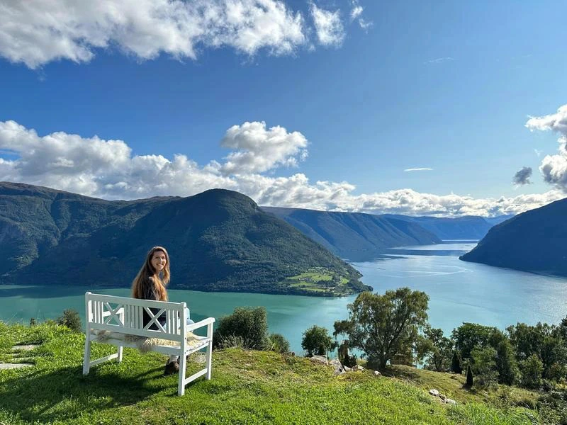 Vrouw zit op een wit bankje op een heuvel en kijkt uit over een fjord met helderblauw water, omringd door groene bergen onder een deels bewolkte blauwe lucht.