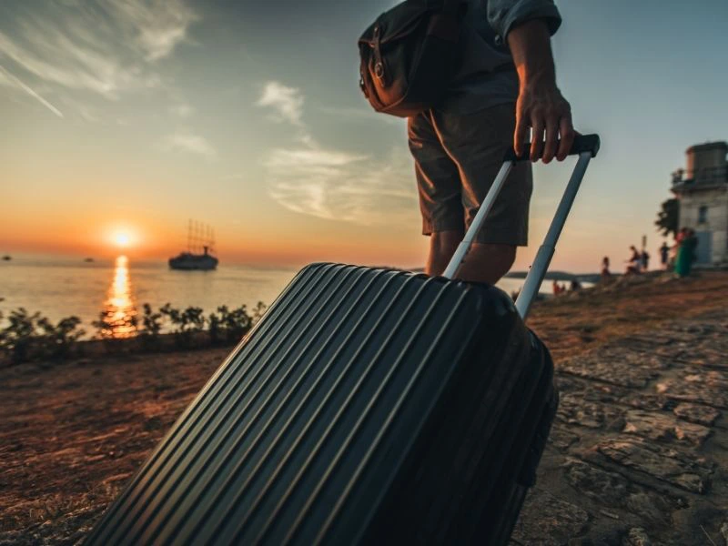 Reiziger met koffer loopt over een stenen pad langs de kust bij zonsondergang, met een schip zichtbaar op zee.