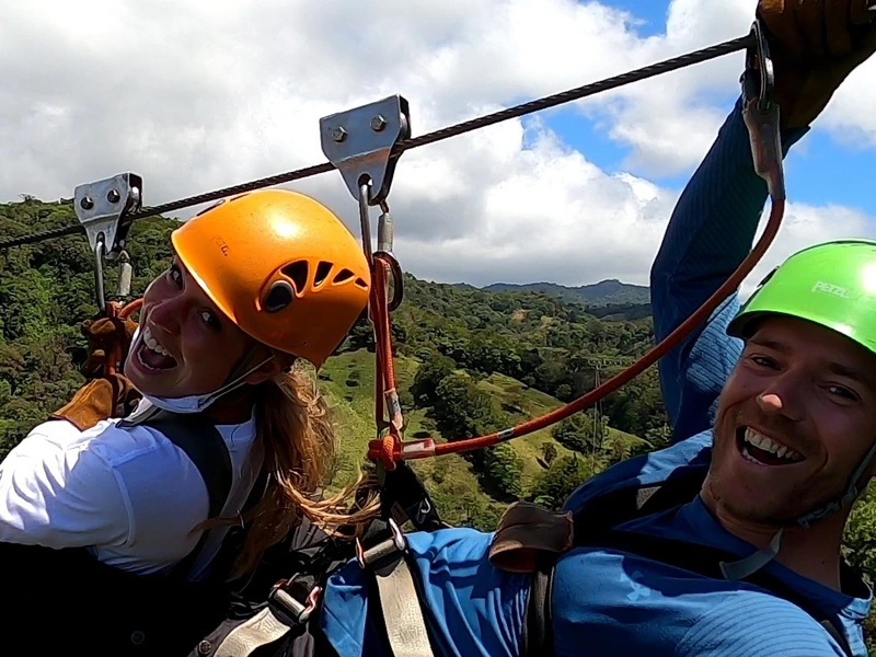 Twee lachende mensen met helmen maken een zipline-tocht boven een groen berglandschap.