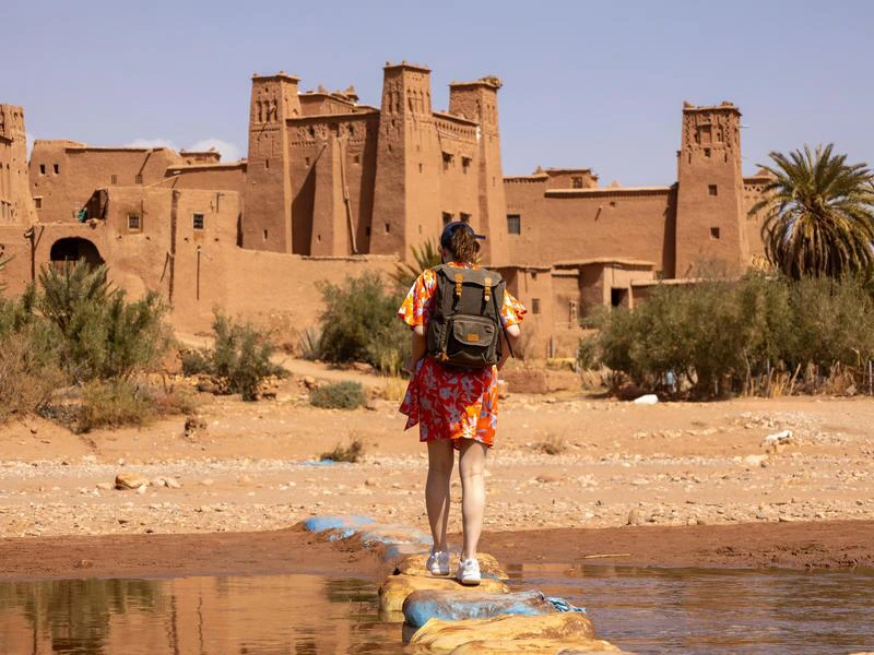 Vrouw met rugzak en rood jurkje steekt een ondiepe rivier over via een stenen pad, met op de achtergrond de lemen vesting Aït-Ben-Haddou in Marokko.