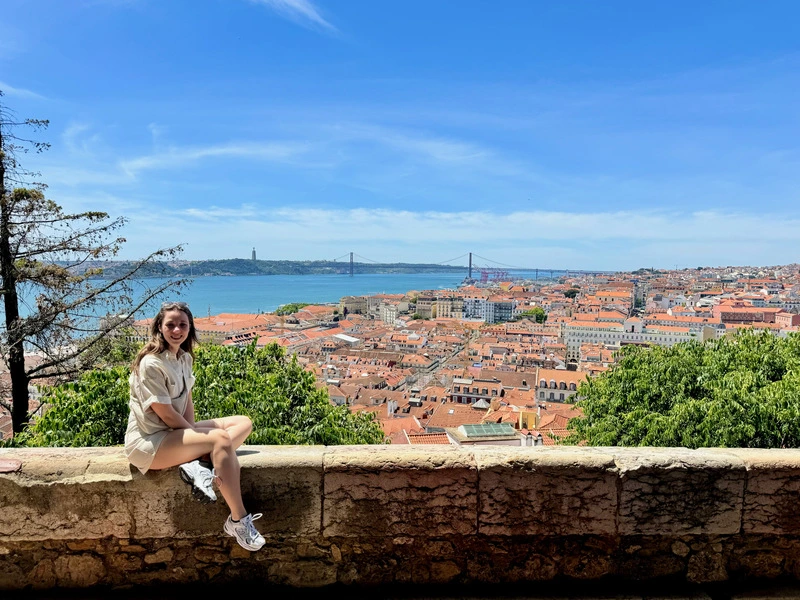 Vrouw zit op een stenen muurtje met uitzicht over de oranje daken van Lissabon en de rivier de Taag, met de beroemde brug Ponte 25 de Abril op de achtergrond.