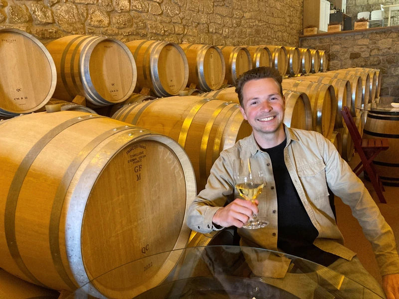 Man zit glimlachend met een glas wijn in de hand in een wijnkelder, omringd door houten vaten.