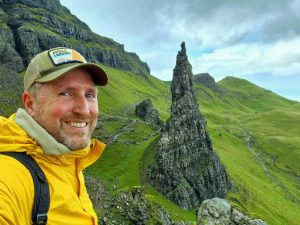 Man in gele jas maakt een selfie met de rotsformatie Old Man of Storr op het groene eiland Skye in Schotland.