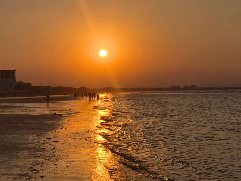 Zonsondergang boven een rustige zee met een gouden gloed op het strand waar mensen langs de waterlijn wandelen.