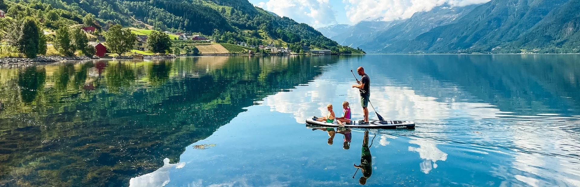 Een persoon staat op een paddleboard met drie kinderen op een spiegelglad meer, omringd door groene bergen en een blauwe lucht met wolken.
