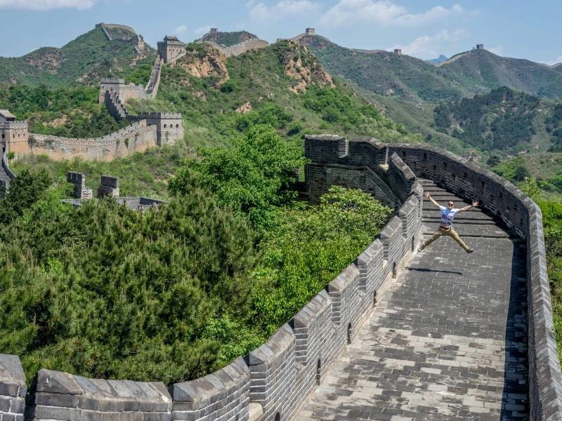 Een persoon springt in de lucht op een stenen pad langs de Chinese Muur, omringd door groene heuvels en wachttorens onder een blauwe lucht.