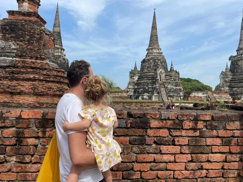Een man draagt een klein meisje op zijn arm terwijl ze samen uitkijken over de oude tempelruïnes van Ayutthaya in Thailand. Voor hen staat een verweerde bakstenen muur, waarachter meerdere grote stoepa’s met spitse torens oprijzen tegen een helderblauwe lucht met lichte wolken.