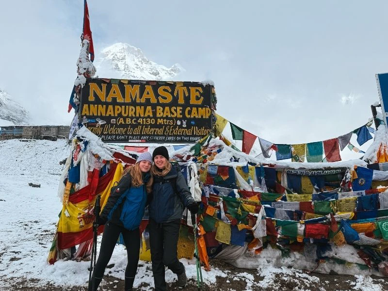 Twee wandelaars poseren lachend bij het bord van Annapurna Base Camp (4.130 meter) in Nepal. Ze dragen warme, waterdichte kleding en wandelstokken, terwijl de grond en omgeving bedekt zijn met sneeuw. Achter hen hangt het bekende zwarte bord met gele letters “Namaste Annapurna Base Camp” en kleurige gebedsvlaggen die in de wind wapperen. Op de achtergrond doemt een besneeuwde bergtop op in de mistige, bewolkte lucht.