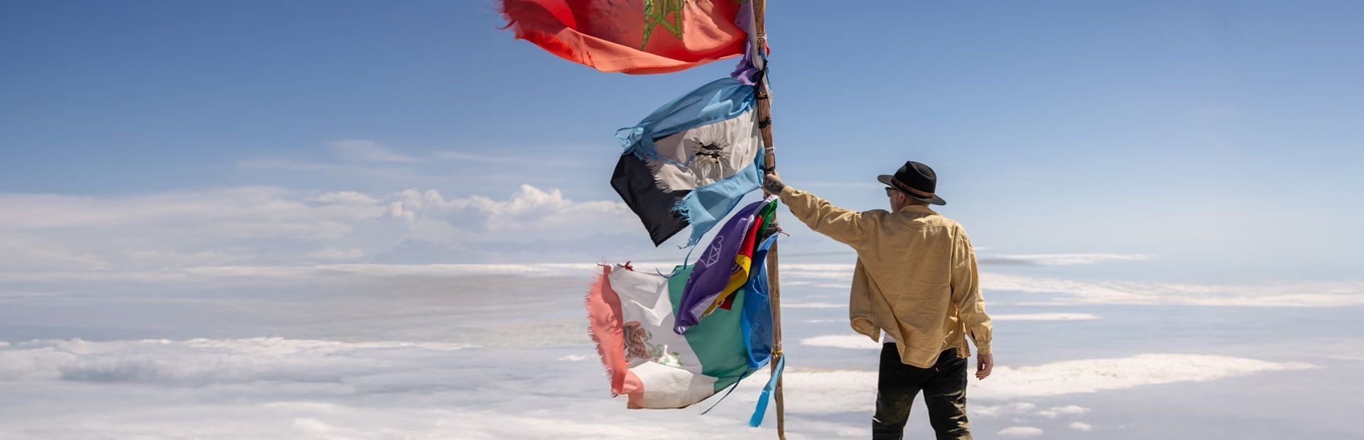 Reiziger op de Zoutvlakten van Boliva bij een vlaggenmast met verschillende landenvlaggen die wapperen in de wind.