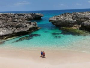 Twee reizigers staand met hun gezicht naar de camera die hangt aan een drone. Ze staan op een wit strand met turquoise water op Sumba, Indonesië. Je ziet ook twee enorme rotsen in de zee die een soort pad vormen voor de zee.