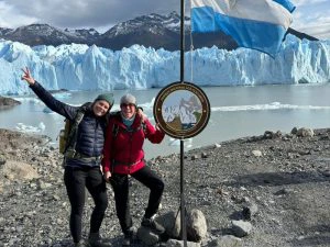 Twee wandelaars poseren lachend naast een bord van Parque Nacional Los Glaciares, met een Argentijnse vlag en de indrukwekkende Perito Moreno-gletsjer op de achtergrond.