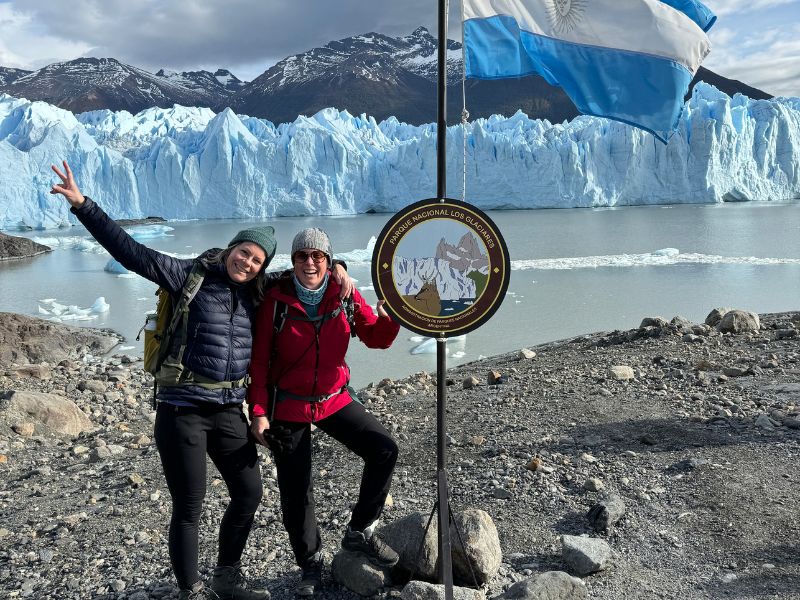 Twee wandelaars poseren lachend naast een bord van Parque Nacional Los Glaciares, met een Argentijnse vlag en de indrukwekkende Perito Moreno-gletsjer op de achtergrond