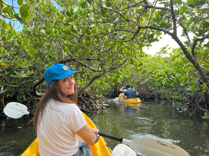 Vrouw in een gele kajak met blauwe pet kijkt achterom en glimlacht, terwijl ze door een smal mangrovebos peddelt. Voor haar vaart nog iemand in een andere kajak