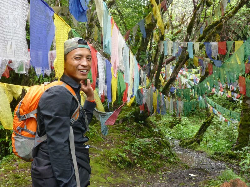 Man met een rugzak glimlacht naar de camera terwijl hij langs kleurrijke gebedsvlaggen loopt in een groene, bosrijke omgeving in Bhutan.