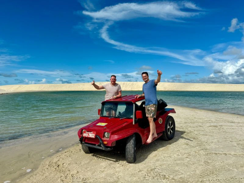 Twee mannen poseren lachend bij een rode buggy op een strand, met een helderblauwe lucht en een lagune op de achtergrond
