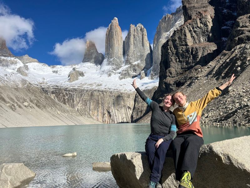 Twee reizigers zitten op een rots bij een helder bergmeer en wijzen lachend naar de iconische granieten torens van Torres del Paine in Patagonië, Chili
