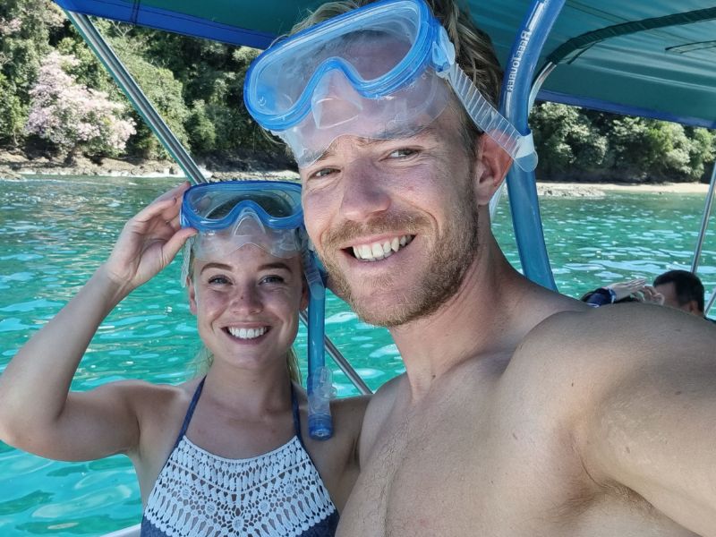Een lachend koppel maakt een selfie op een boot, beide dragen snorkelmaskers op hun hoofd. Het heldere turquoise water en de groene kustlijn zijn op de achtergrond te zien