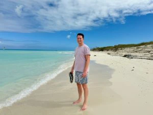 Man staat op een wit zandstrand aan een turquoise zee, met slippers in de hand en een blauwe lucht met wat wolken boven hem