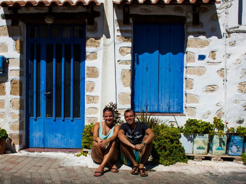 Koppel zit lachend op straat voor een wit stenen huis met felblauwe deur en luiken, omringd door planten