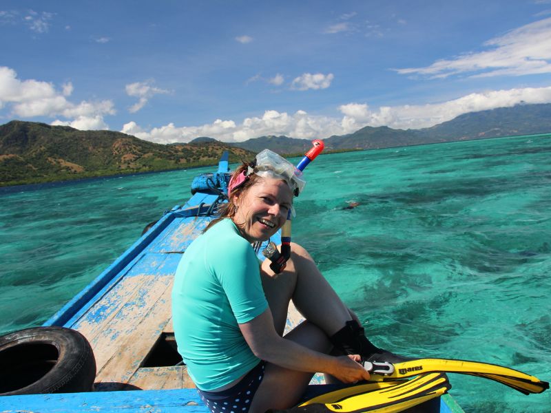 Een vrouw zit lachend op de rand van een houten boot, met snorkelmasker en vinnen, klaar om het heldere turquoise water in te gaan. Op de achtergrond zijn groene heuvels en een blauwe lucht te zien
