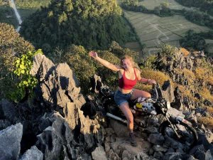Vrouw poseert vrolijk op haar motor op een rotsachtige bergtop in Laos, met uitzicht op groene rijstvelden en heuvels in de verte