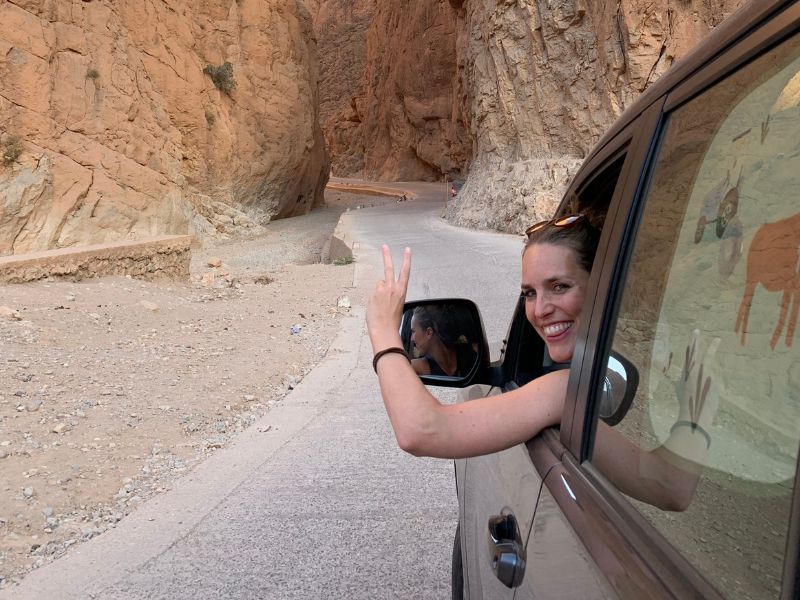 Vrouw steekt haar hand met een peace-teken uit het raam van een auto terwijl ze door een smalle kloof in een woestijnachtig landschap rijdt