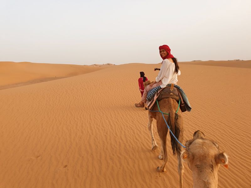 Vrouw met rode hoofddoek rijdt op een kameel door een uitgestrekte woestijn met zandduinen
