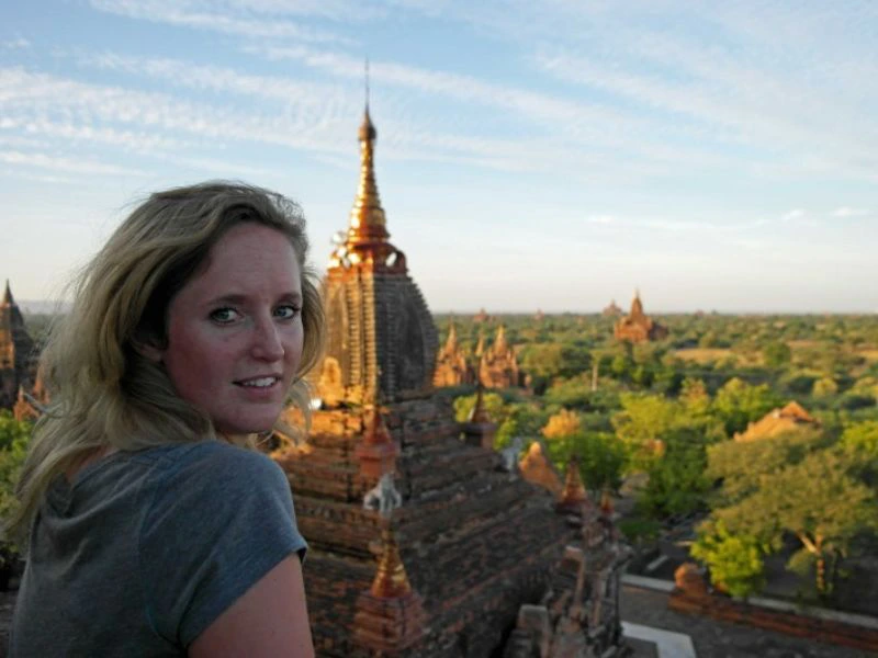 Vrouw kijkt over haar schouder vanaf een tempel in Bagan, Myanmar, met uitzicht op eeuwenoude pagodes en groene vlaktes in de avondzon.