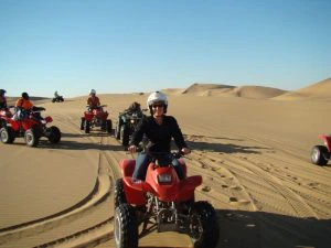 Een groep mensen rijdt op quads door een uitgestrekte woestijn met hoge zandduinen, met een helderblauwe lucht op de achtergrond
