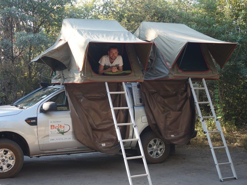 Man ligt lachend in een daktent op een 4x4-camper. Geparkeerd in een bosrijke omgeving
