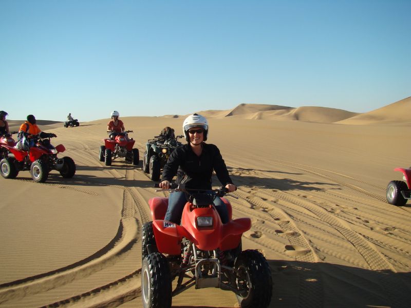Een groep mensen rijdt op quads door een uitgestrekte woestijn met hoge zandduinen, met een helderblauwe lucht op de achtergrond